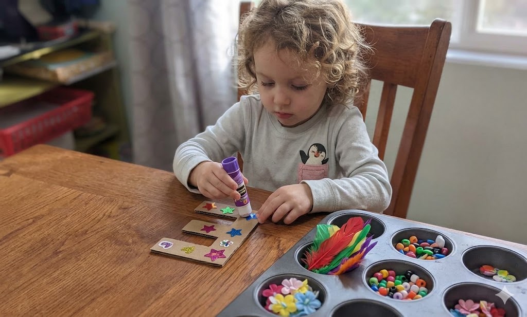 A child sitting at a table gluing bright colored stars onto a cardboard letter 'E'. A muffin tin filled with beads, feather and other craft supplies is on the table next to her.